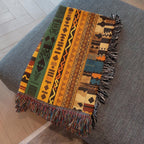 Kök Stüdyo woven throw blanket with Ndebele-inspired geometric patterns in green, yellow, orange, and blue. Features fringe detail.