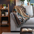 Kök Stüdyo Ndebele-inspired woven throw blanket with geometric patterns in tan, blue, and rust on a grey sofa.